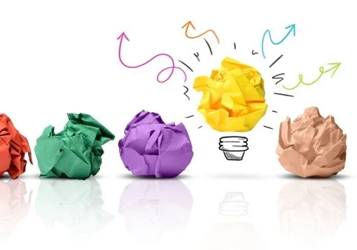 Colourful rolled up balls of paper laid out on a white table with one turning into a lightbulb.