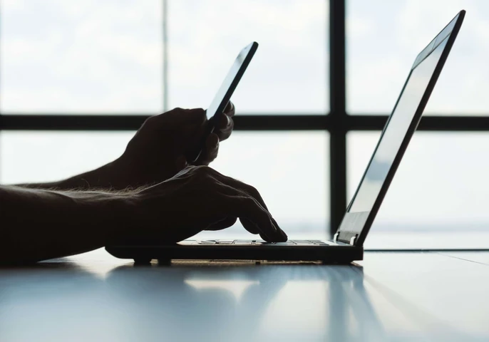 A person working on a laptop while holding a smartphone