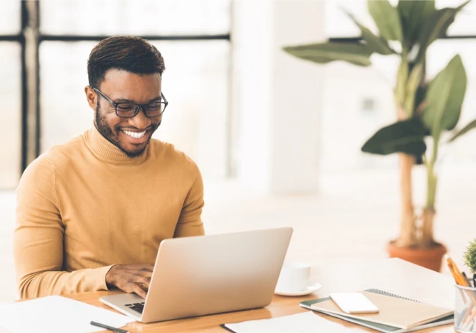 A man in a beige turtleneck sitting at a desk and working on a laptop.