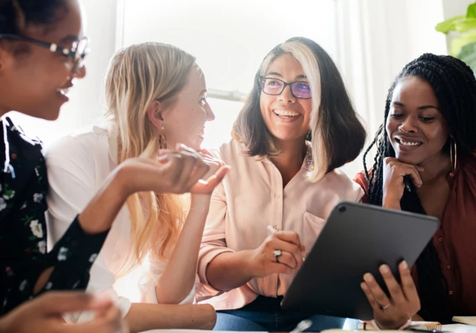 A group of four women sitting at a desk, invested in a discussion around a tablet.