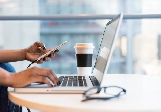 Person at a desk typing on a laptop keyboard and holding a smartphone