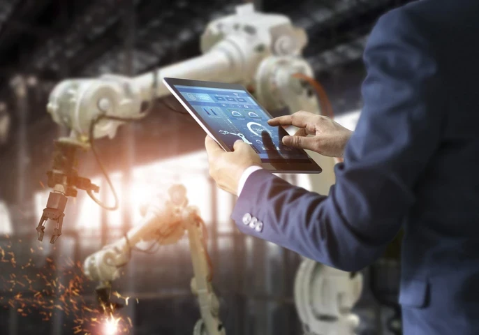 Man in a suit tapping on a tablet while a manufacturing robot works in the background.