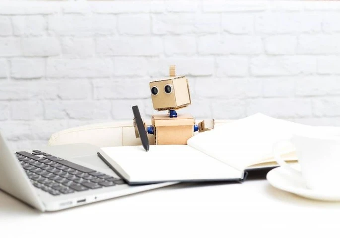 Toy robot pretending to write in a notebook while looking at a laptop