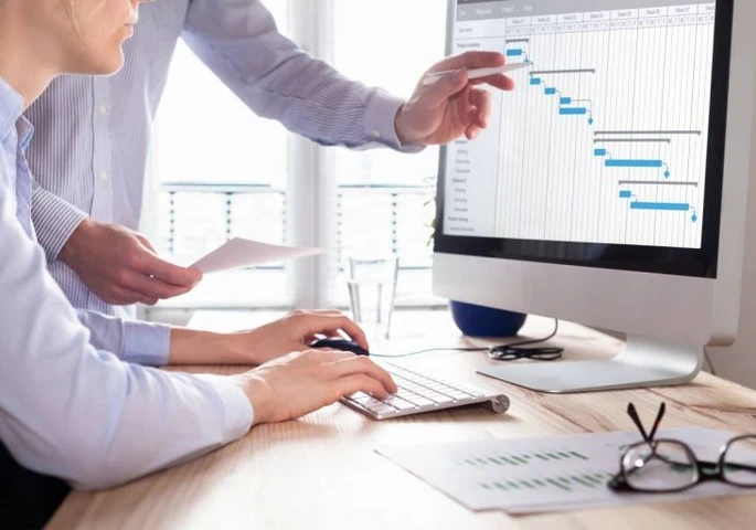 Two businesspeople standing and sitting in front of a desktop computer viewing analytics on a screen.