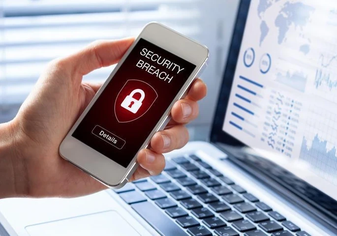 Person holding a smartphone with the words "Security Breach" and a lock in front of a laptop displaying graphs.