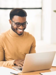 A man in a beige turtleneck sitting at a desk and working on a laptop.
