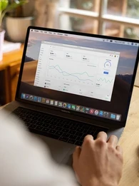 Person sitting at a desk working on a laptop with graphs on the screen.