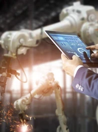 Man in a suit tapping on a tablet while a manufacturing robot works in the background.