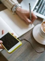 person working on a smartphone, making notes in a book with a coffee and laptop beside them