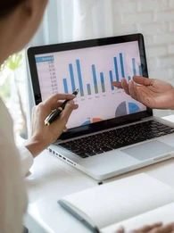 Two people sitting at a desk pointing at a laptop screen with graphs on it.