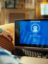 A woman sitting on a couch with a virtual customer support agent on a laptop.