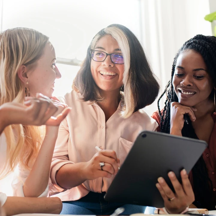 A group of four women sitting at a desk, invested in a discussion around a tablet.