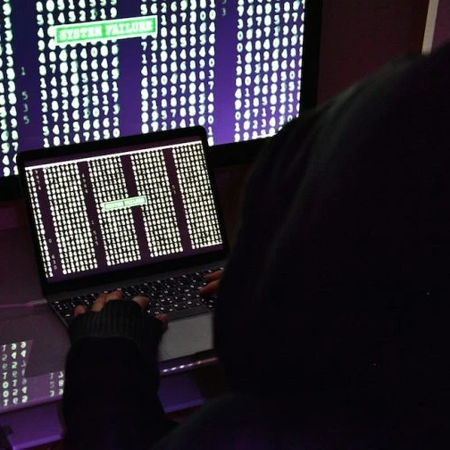 Hacker in a black hoodie sitting in front of two computer screens with lines of code.