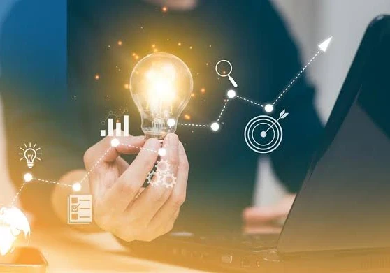 A person holding up a lightbulb while sitting in front of a laptop with icons floating around.